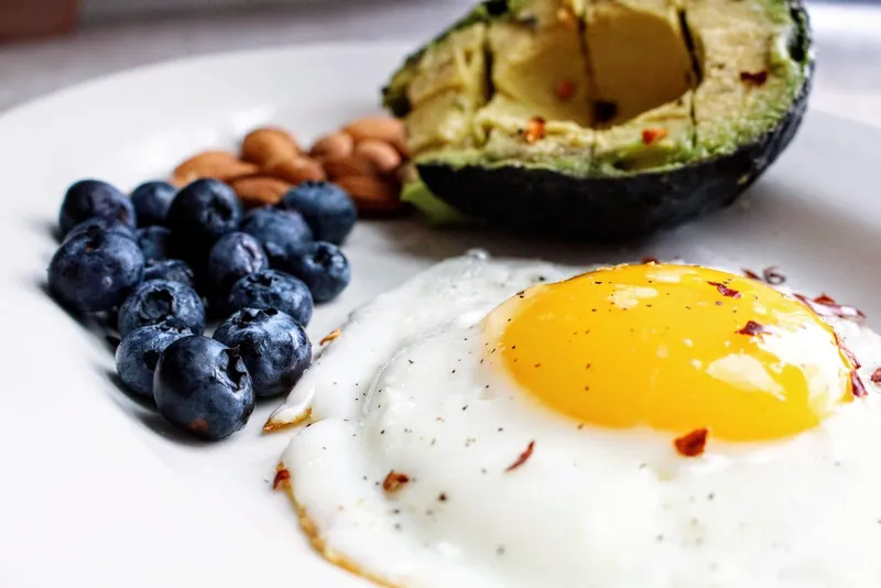 Desayuno keto con huevo frito, aguacate, arándanos y almendras sobre un plato claro